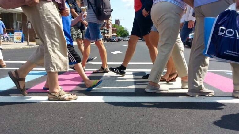 People, obsured from the waist up, are seen walking across a crosswalk that is painted with the colours of the transgender flag.