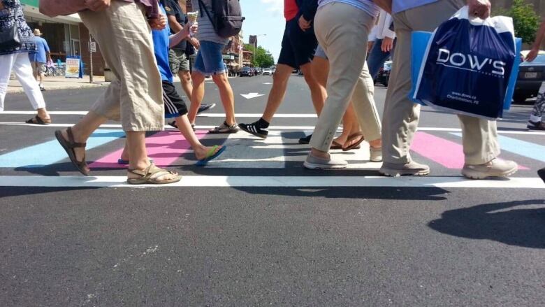 People, obsured from the waist up, are seen walking across a crosswalk that is painted with the colours of the transgender flag.