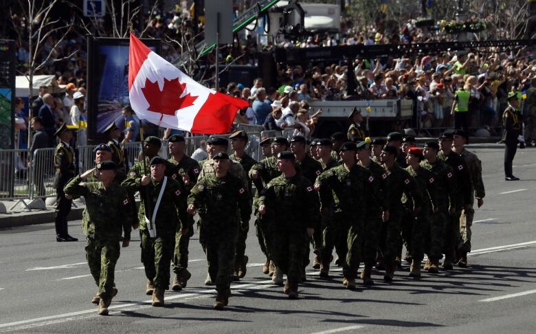 A group of soldiers march down a street on a sunny day with crowds and a Canadian flag in the background.