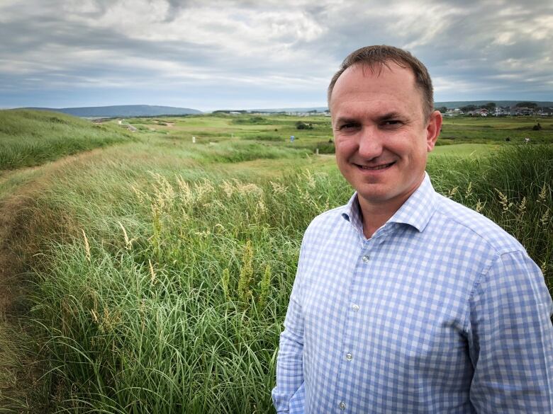 A mman stands in the foreground with dune grasses and a golf course in the background.