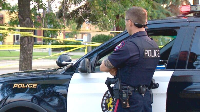 A Calgary police officer stands beside a police car near yellow crime scene tape.