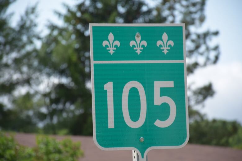 A green highway sign labeled '105.' There are fleurs-de-lis across the top. The background is blurred but there is a beige road, green trees, and a blue sky.