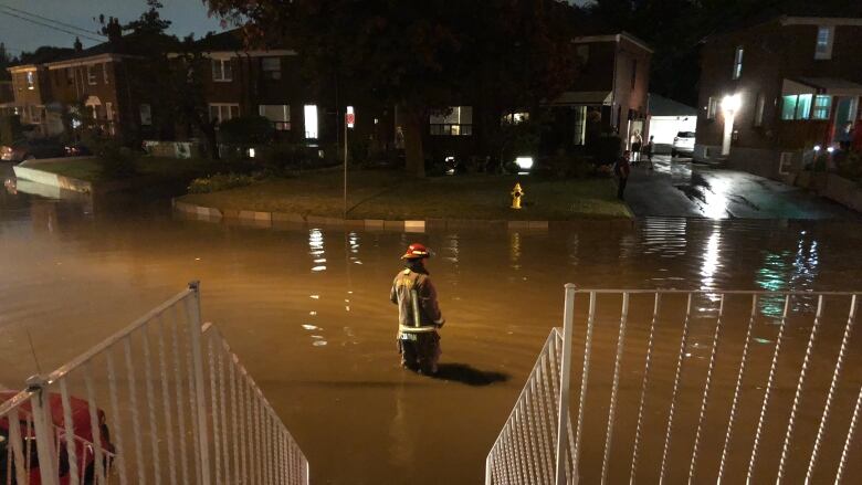 At night, a firefighter stands in the middle of a residential city street, up to his knees in floodwater.