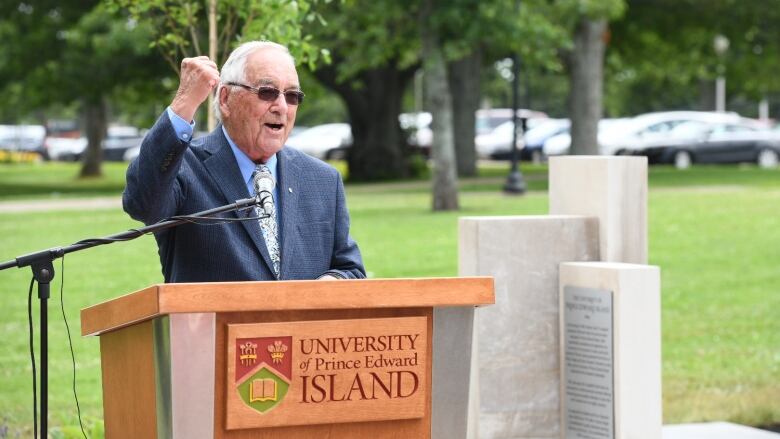 Smiling man in business suit and sunglasses raises his fist in celebration as he stansd outside behind a University of Prince Edward Island podium. 