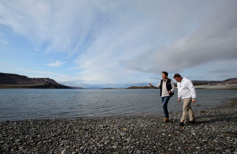 Two men walk on a gravel beach in the High Arctic with tundra covered mountains in the background. 