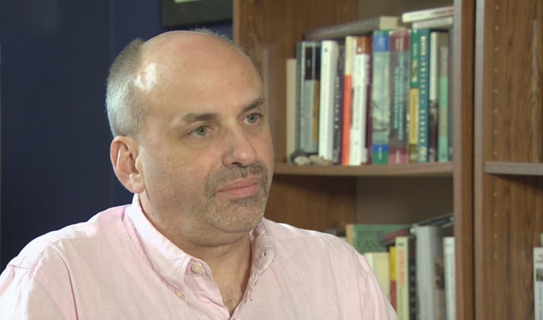 A man with short gray hair and a starting goatee and wearing a pale coloured button down dress shirt with the first button undone looks a bit to the right of the camera. In the background is a medium brown wooden bookshelf filled with books and a navy blue wall.