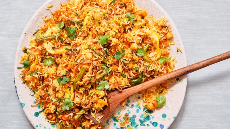 Overhead shot of a serving plate of biryani on a grey surface. The rice is golden and green beans, carrots, fried onions and cilantro are visible on top. A wooden spoon sits in the rice. 