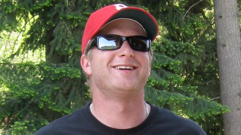 A family photo shows a white man in his 30s with short hair smiling in front of an evergreen tree. He is wearing black sunglasses, a red baseball cap and a black T-shirt.