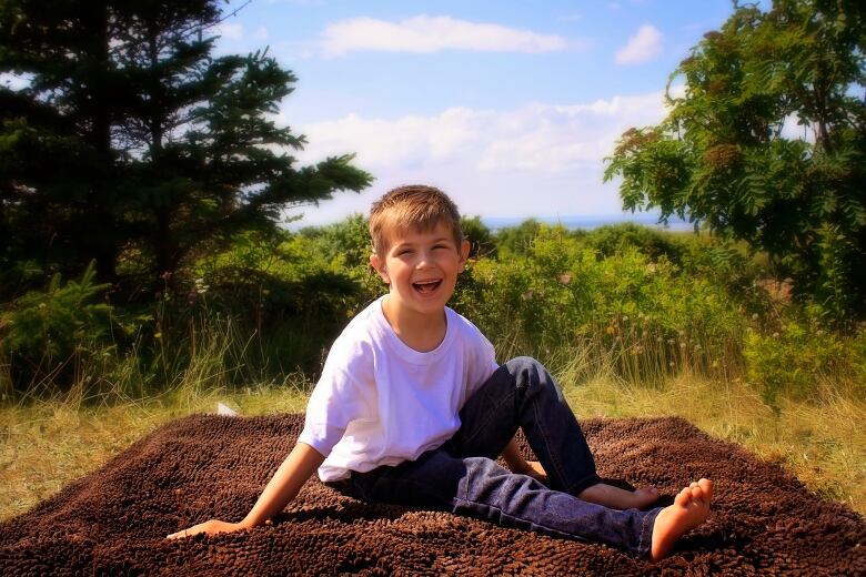 A boy wearing a white shirt with light brown hair sitting on a blanket outside in the summer. He is smiling.