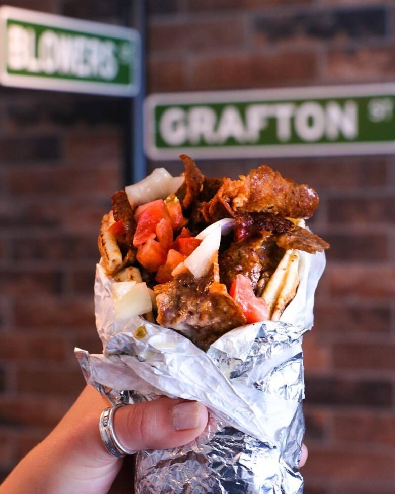 A hand holds a tin foil-wrapped donair with pits, meat, tomatoes, onion and donair sauce.