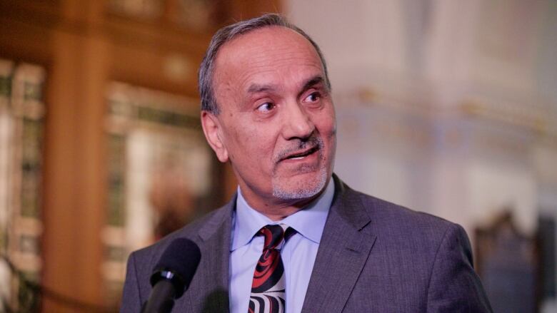 An older South Asian man wearing a patterned tie looks away from a mic while standing in an old building.