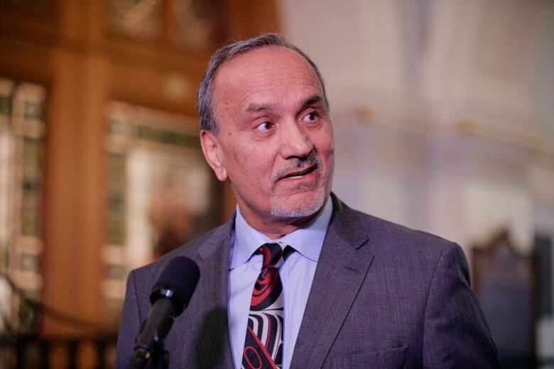 An older South Asian man wearing a patterned tie looks away from a mic while standing in an old building.