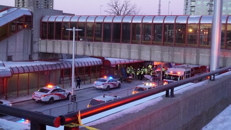 Police cars line up beside a transit stop, while people in florescent jackets surround a crash site