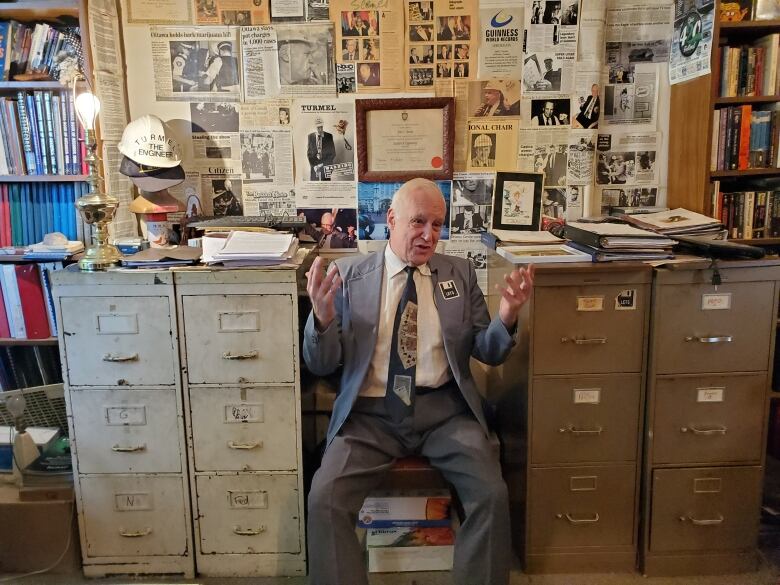 A person sits between a bunch of filing cabinets in an office with clippings all over the wall.