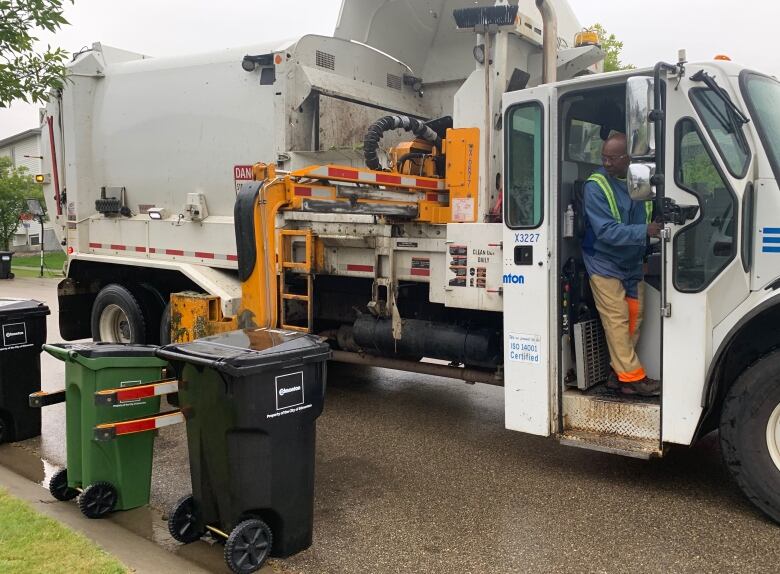 A man dressed in blue stands at the door of a waste collection truck. Waste bins sit along the curb. 