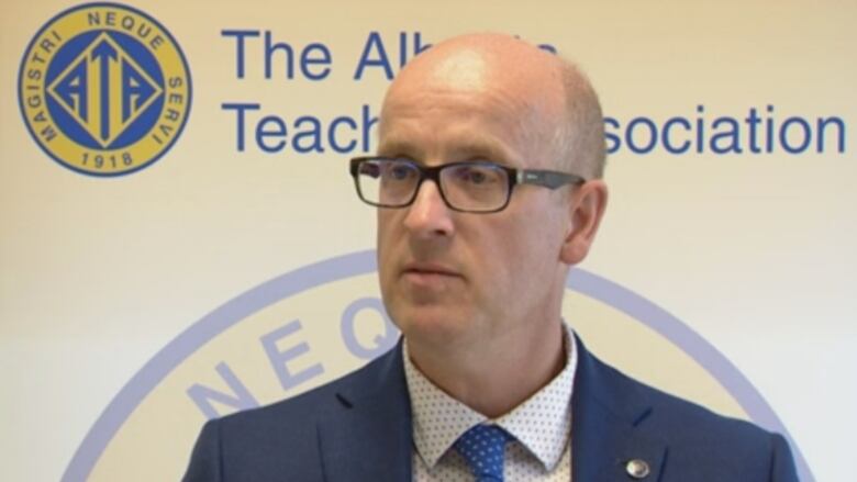 A man wearing a suit and glasses stands in front of a backdrop that reads, Alberta Teachers' Association.