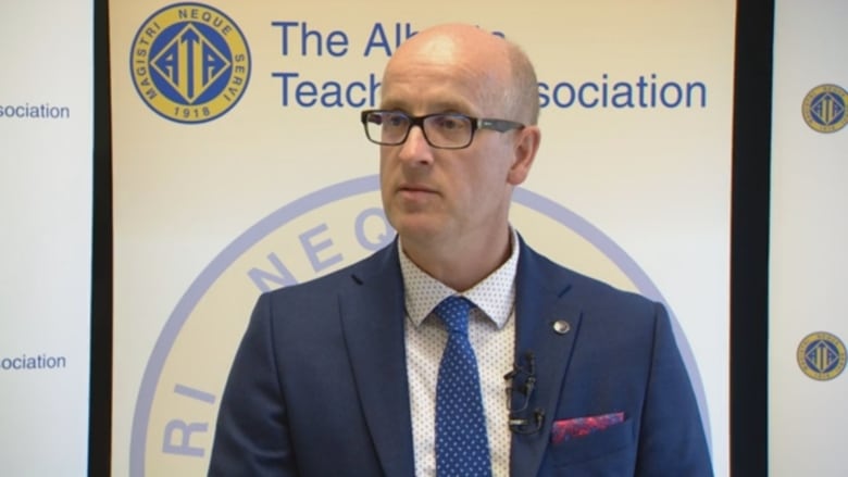 A man wearing a suit and glasses stands in front of a backdrop that reads, Alberta Teachers' Association.