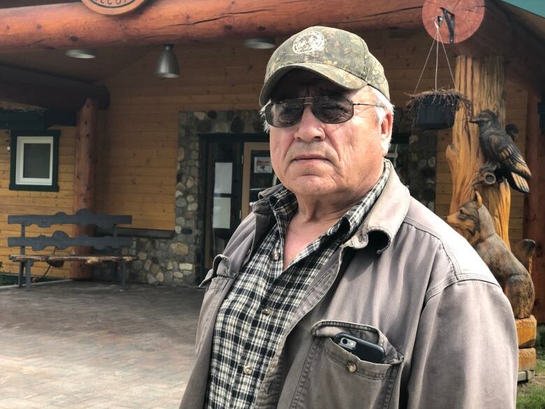 Man wearing a hat and sunglasses standing infront of a log cabin.