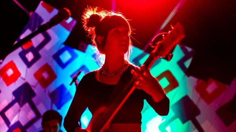 A woman with dark hair in a bun is holding a guitar on stage. She is wearing a black long sleeve shirt and gold chain necklace. She is looking into the distance to her left, and stands in front of a background with squares of different colours and widths.