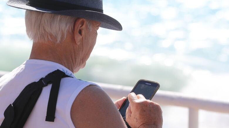 An elderly man scrolls on a mobile phone while sitting on a bench near a beachfront. He sits next to an elderly woman. 