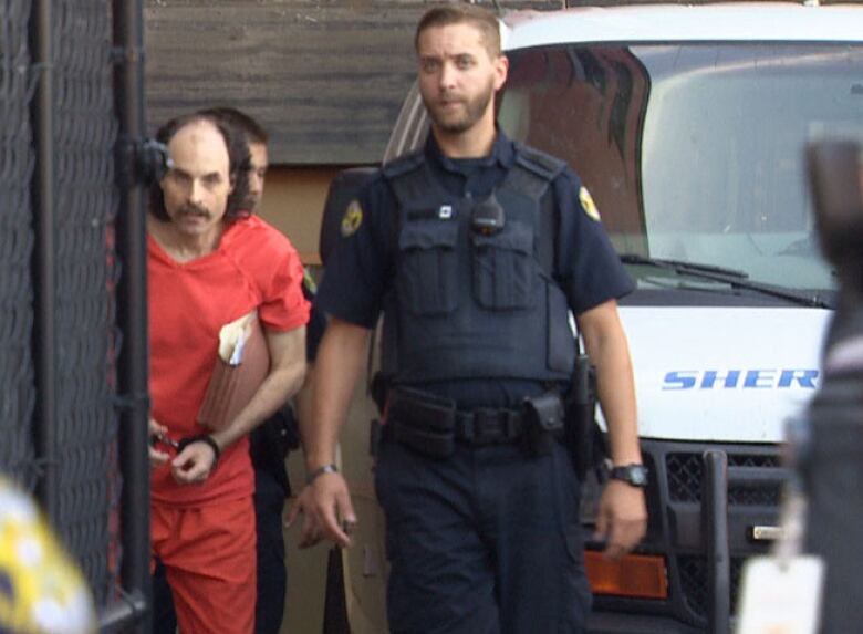 A man in an orange jumpsuit is led by uniformed peace sheriffs.