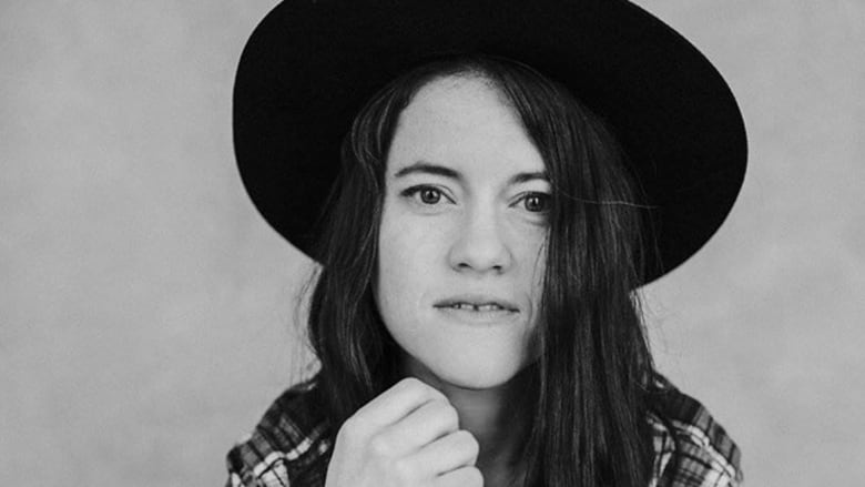 A woman with long black hair and a wide-brimmed hat and wearing a checked shirt looks into the camera in this black and white photograph.