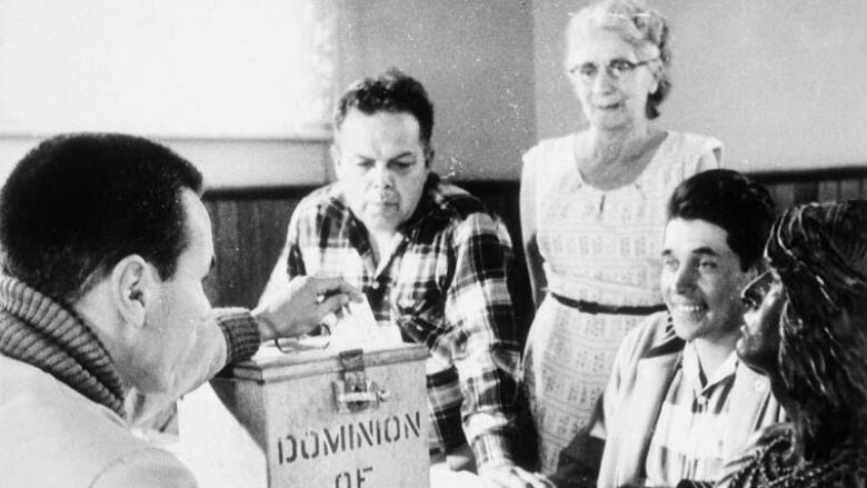 Black-and-white photo shows a man placing a ballot into a Dominion of Canada voting box while other individuals look on. 