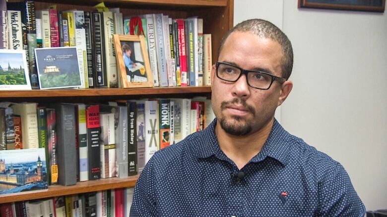 A man with glasses and a goatee sits in front of a bookcase. 