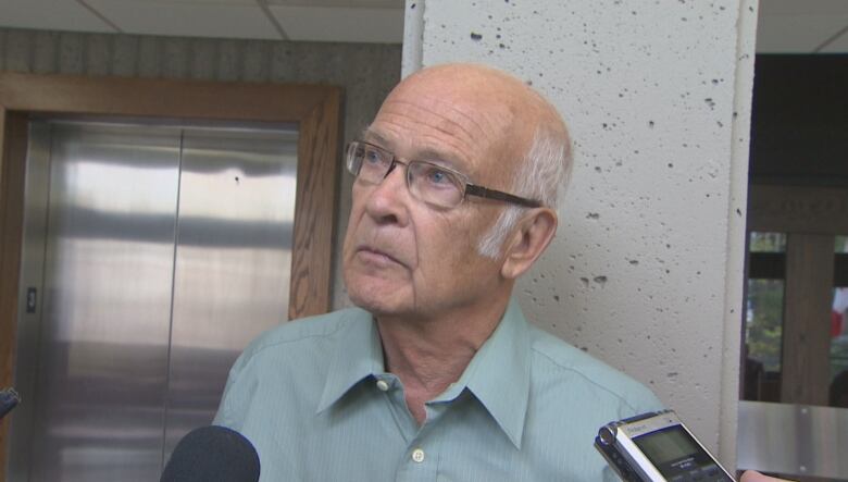 An old, balding white man is seen being interviewed by reporters. He's wearing thin wire-rim glasses and a short sleeved, green button up shirt.