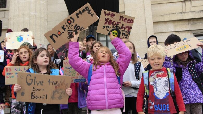 Elementary-aged students in bright clothing stand at  the Manitoba Legislature holding signs with messages on climate change.