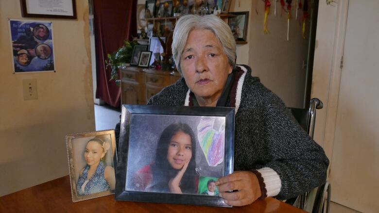 A woman sits at a table and holds up two photos that show teenage girls. 