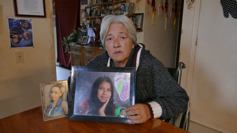 A woman sits at a table and holds up two photos that show teenage girls. 