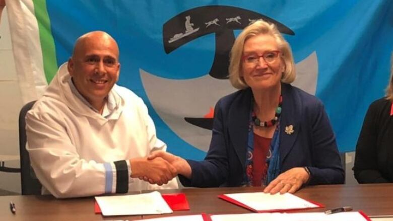 A man in a white top shakes hands with a woman in a blue jacket. They are sitting at a table with papers on it.