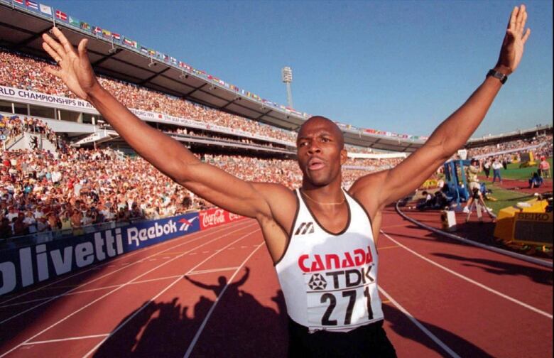 A sprinter stands on a track with both of his arms raised. Thousands of people are in the stands behind him.