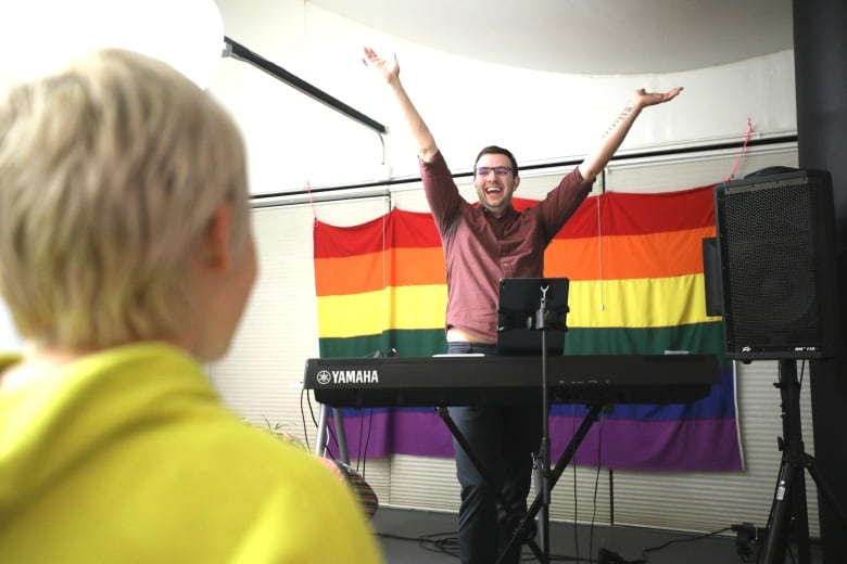 A bearded man stands with his arms extended at the front of a classroom, with a student's back in the foreground and a rainbow flag in the background. 