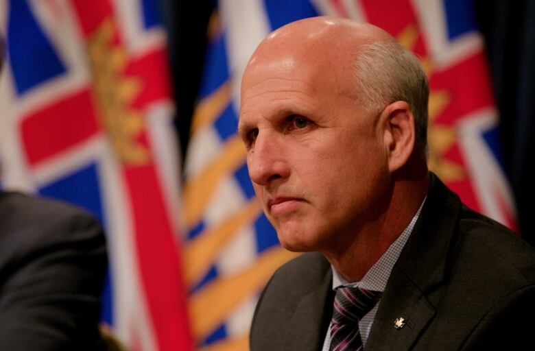 A bald white man looks on at a press conference, with two B.C. flags visible behind him.