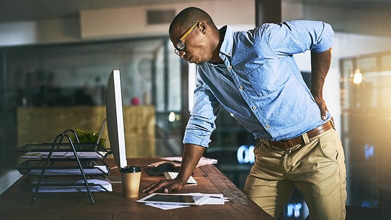 A man hunches over in visible pain, clutching his back while stabilizing his body against a desk.