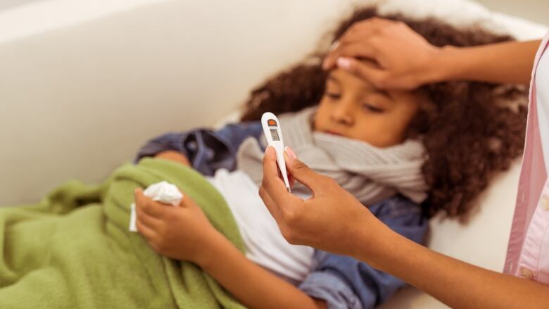 A parent places their hand on the forehead of a young child while looking at a thermometer in their other hand.