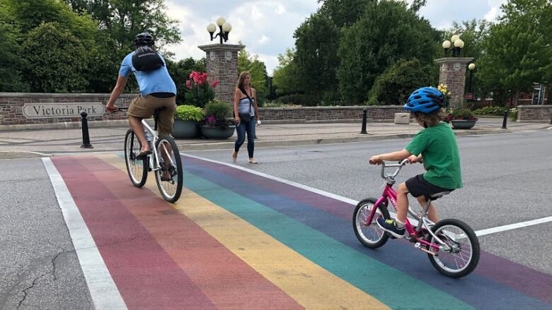 An adult and child cycle over a rainbow crossing into a park.