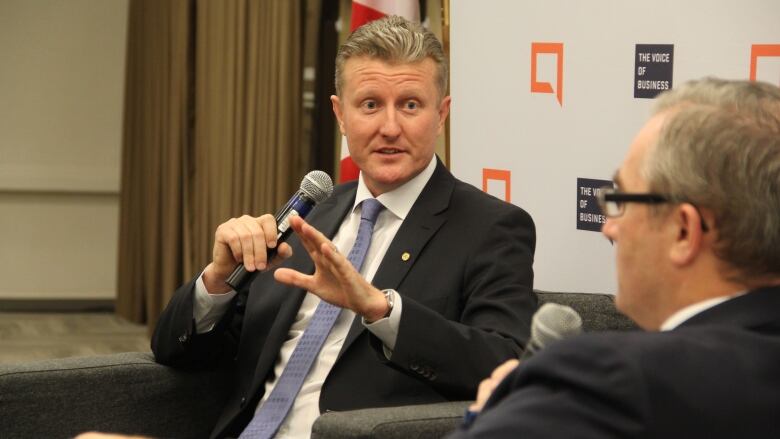 A man in a suit gestures with his left hand while holding a microphone in his right hand.