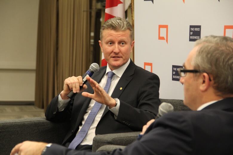 A man in a suit gestures with his left hand while holding a microphone in his right hand.