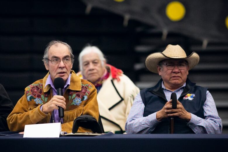 A man, sitting at a table, speaks into a microphone. While another man, sitting beside him and wearing a cowboy hat, looks on. 