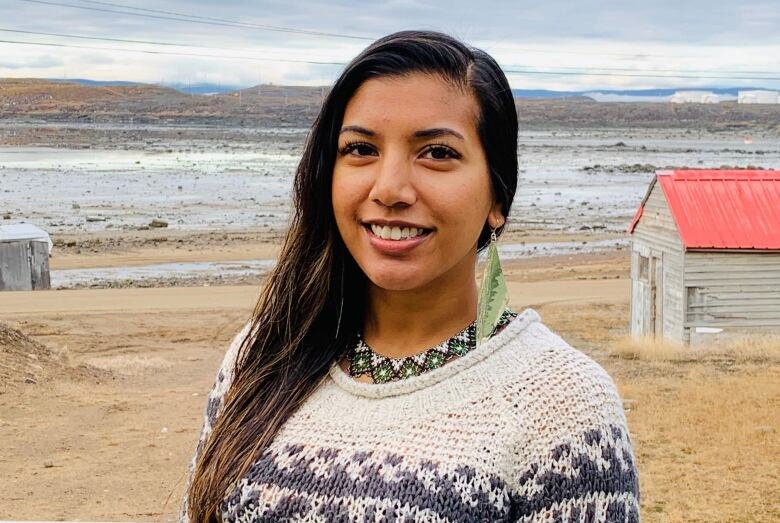 A woman wearing a knitted sweater and long, dangling earrings poses for a photo by the beach in Iqaluit. 