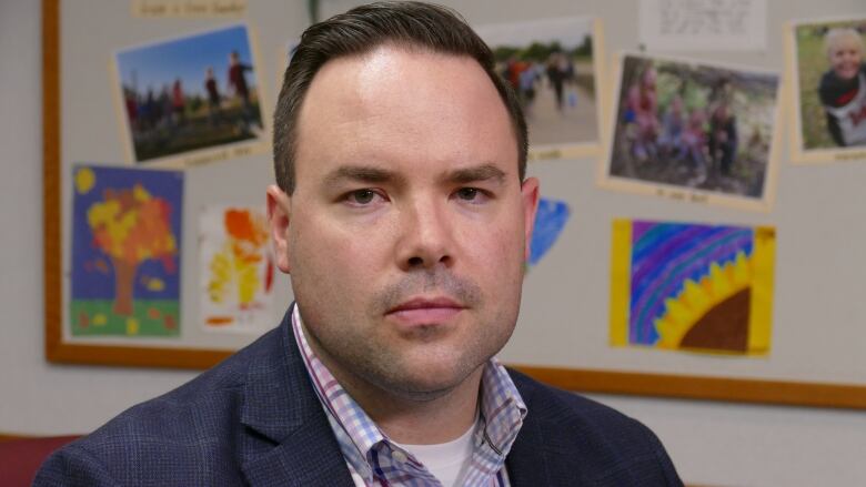 A man in a suit looks serious in front of a wall covered with children's artwork.