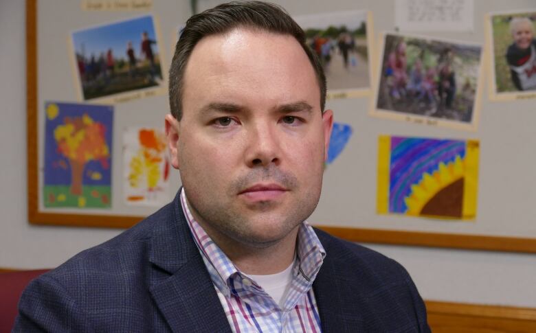 A man in a suit looks serious in front of a wall covered with children's artwork.