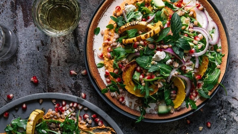 A big dinner plate with salad, sitting on a dark grey surface. The salad is made of slices of delicata squash, red onion, pomegranate arils, greens and herbs. A glass of white wine sits next to it. 