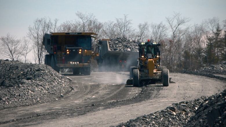 Dumptrucks and a tractor drive along an industrial road, lined with rocks.