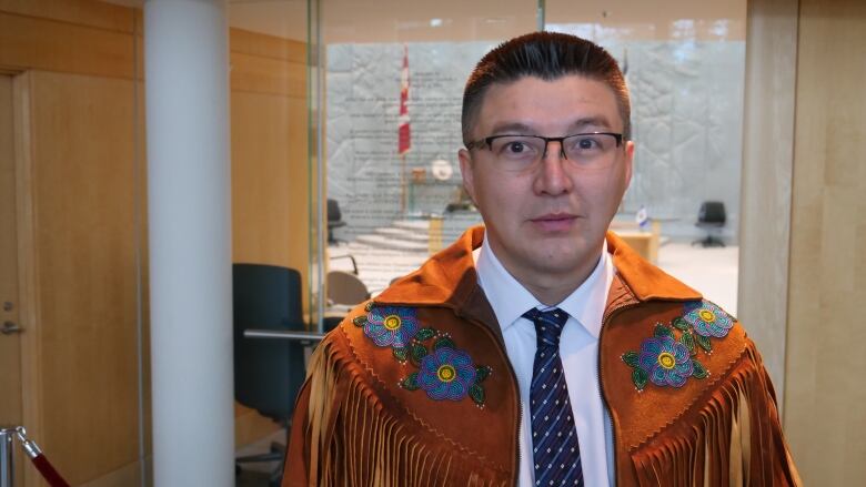 A man wearing a collared shirt with tie and over top a tan moose hide beaded jacket