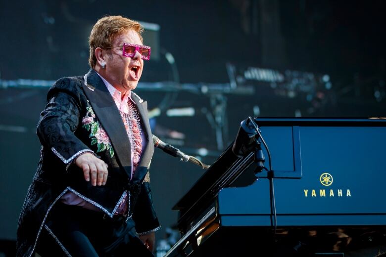 A light-haired man in a dark suit rimmed with sparkles sings while standing by a black piano.