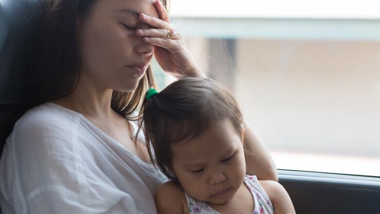 A mother holds her head in her hand while her child looks at an electronic screen.